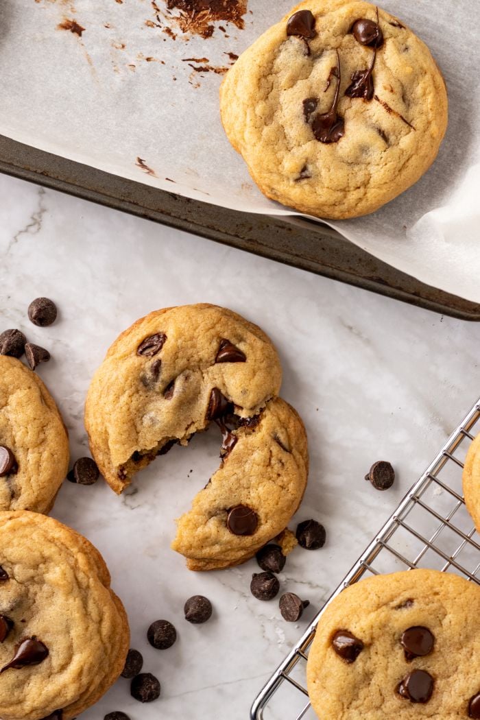 chocolate chip cookies are shown on a baking sheet, a cooling rack, and the countertop in a top down shot